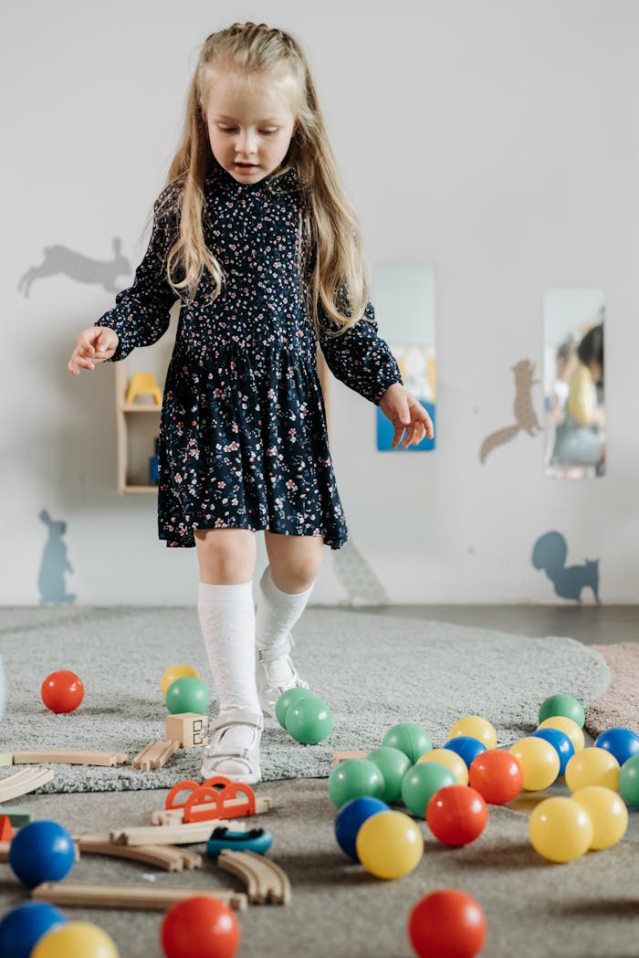 Paslaugos A preschool girl enjoys playtime indoors with colorful toys and a floral dress.